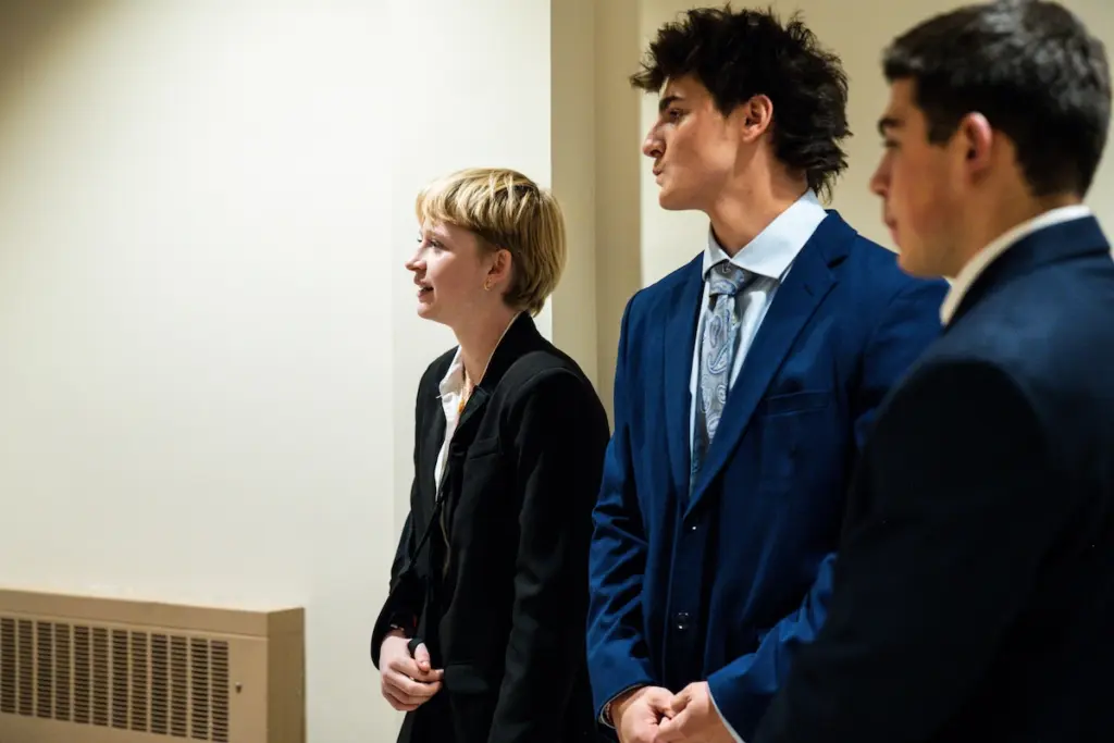 Three pre-law students in business attire stand indoors, facing left. One person has short blonde hair, and the other two have dark hair. They appear attentive and are possibly in a professional or formal setting.