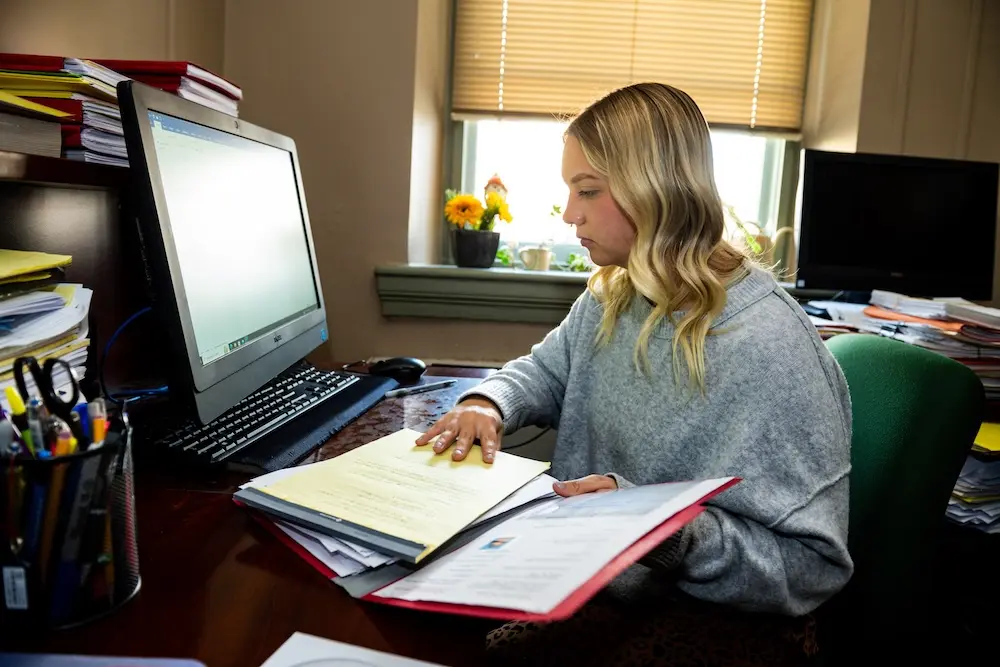 A woman with blonde hair, wearing a gray sweater, sits at a desk with a computer. She is reviewing documents in a folder. The room is well-lit with a window in the background, with sunflowers on the windowsill and stacks of papers nearby.