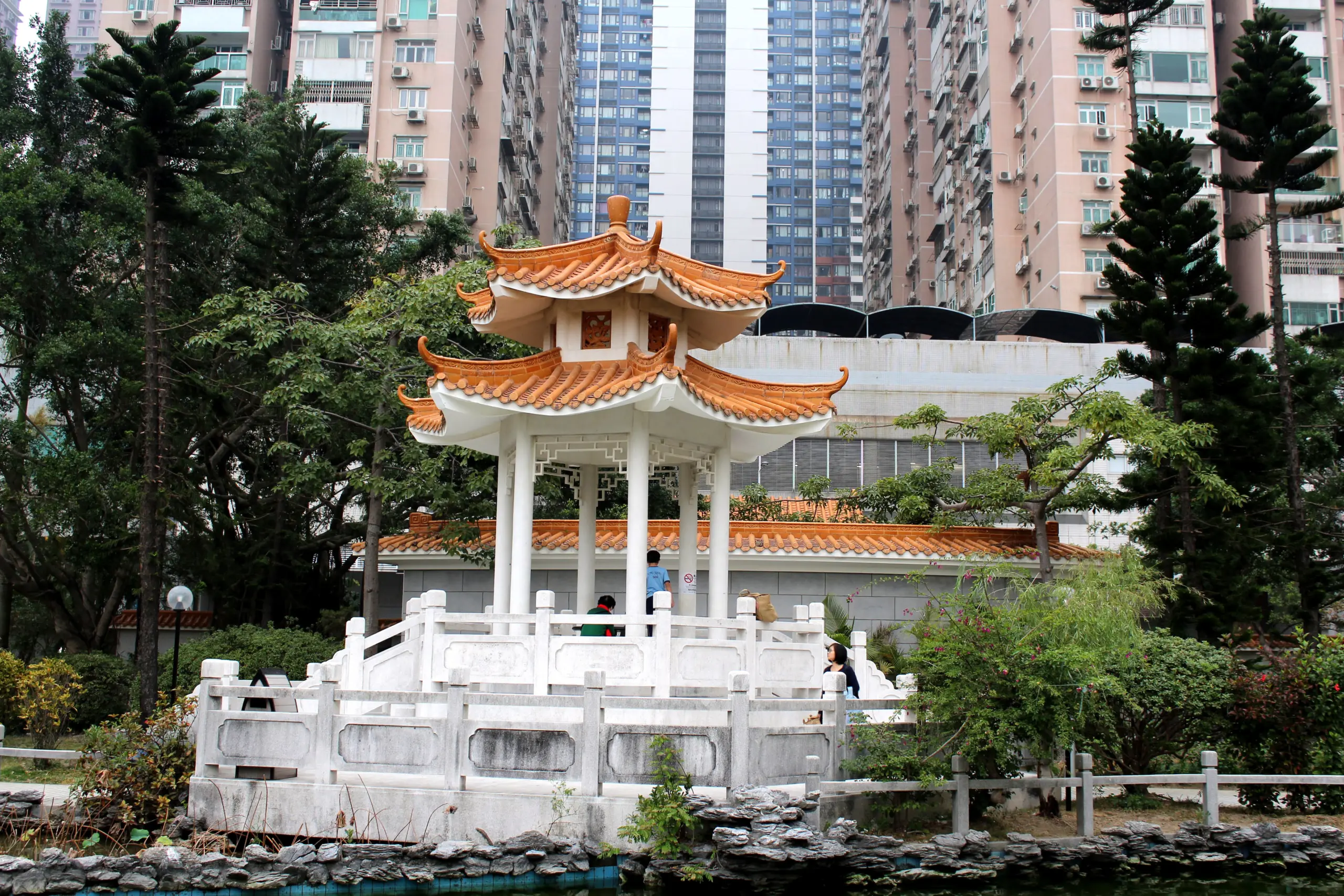 A traditional Chinese pavilion with an orange, tiled roof stands gracefully in a garden of green foliage, complemented by a small pond. Behind the pavilion, towering modern apartment buildings create a striking contrast, symbolizing the harmony of history and progress akin to applied linguistics.