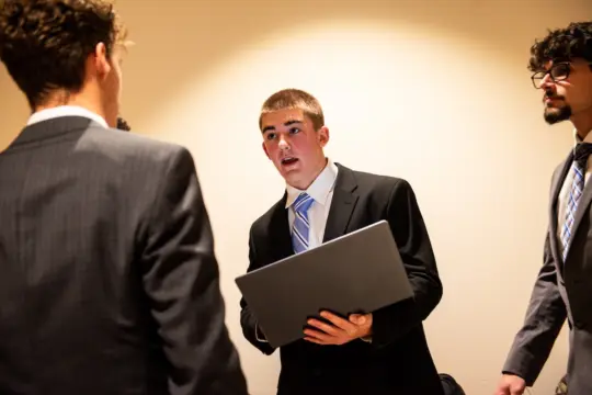 A young man in a suit holds a laptop, engaged in conversation with two other men in suits. The setting appears to be professional or business-related, with a neutral beige background.