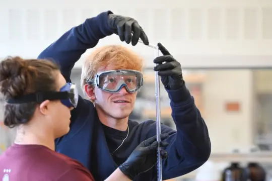 Two students in a science lab. A person with blonde hair and safety goggles holds a tall measuring instrument, while a second person observes. Both wear protective gloves and eyewear, focusing on the task.