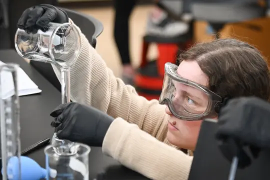 A person wearing protective goggles and gloves carefully pours liquid from one beaker into another in a laboratory setting. They're focused on their task, with additional lab equipment visible in the background.
