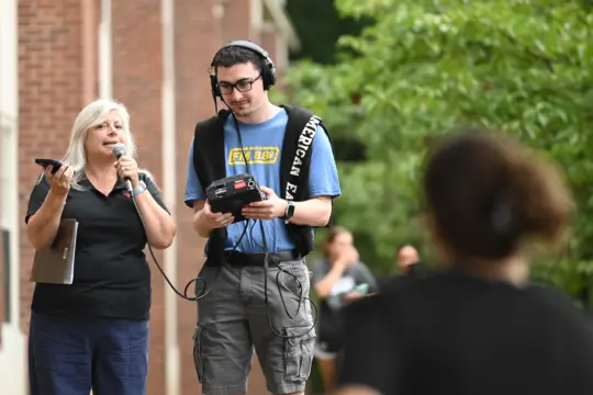 A woman with a phone speaks into a microphone, embodying the essence of Communication Arts. Beside her, a man with headphones skillfully manages audio equipment. They're outdoors near a brick building, while a tree adds to the serene backdrop and a blurred figure stands in the foreground.