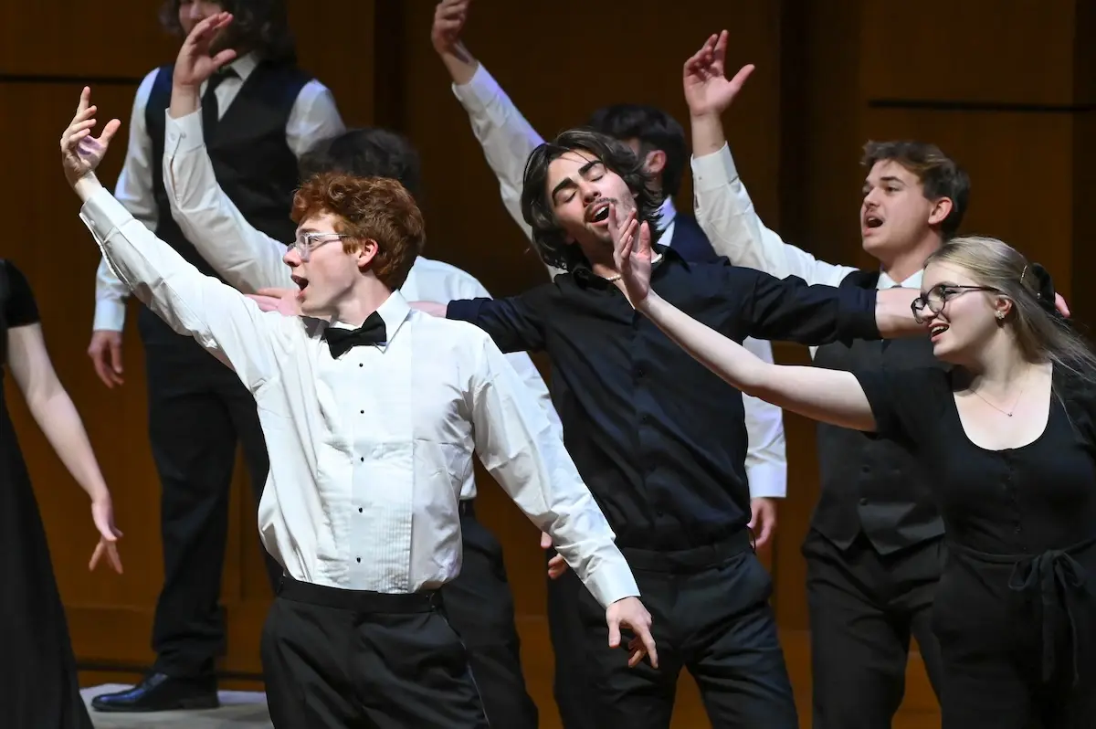 A group of young performers on stage, energetically singing and gesturing with their arms raised. They are dressed in formal attire, including white shirts, black vests, and dresses, and are mid-performance with expressions of enthusiasm.