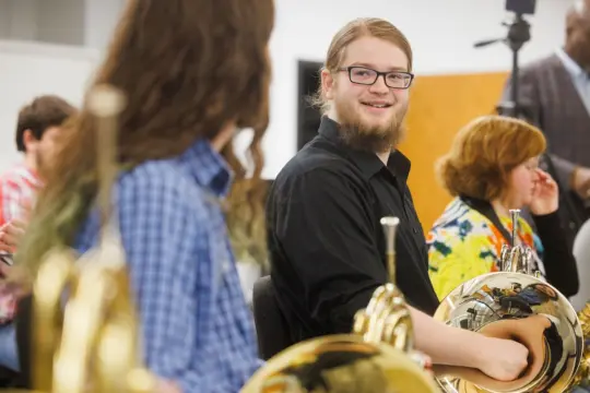 A young man with glasses, wearing a black shirt, is smiling and holding a French horn. He is sitting among others in a music class or rehearsal room. Several people are visible in the background, each holding musical instruments.