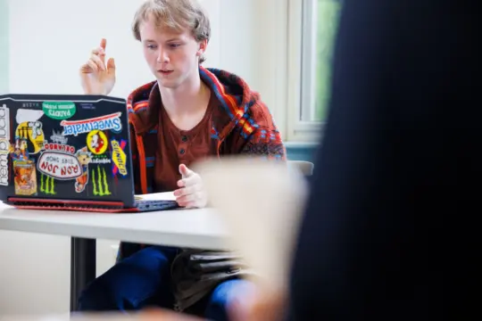 A person sits at a desk, engaged in conversation, with a focused expression. They use a laptop adorned with colorful stickers. A window is visible in the background, and another person is partially seen in the foreground.