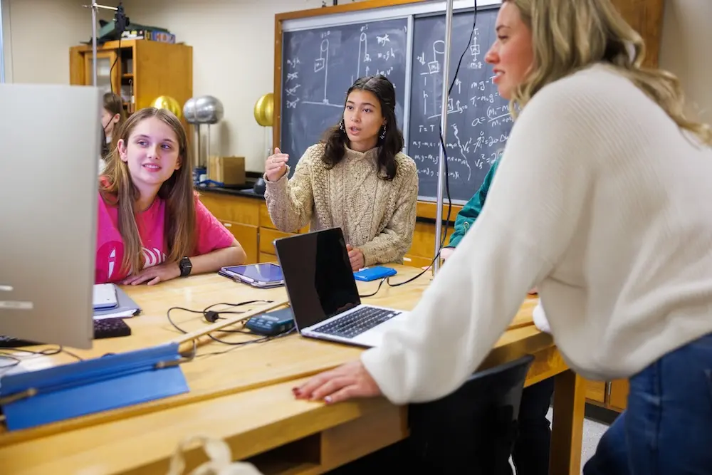 A group of students, including three women, are engaged in a discussion around a table with laptops and notebooks. A blackboard with equations is in the background. They appear to be in a classroom or lab setting.