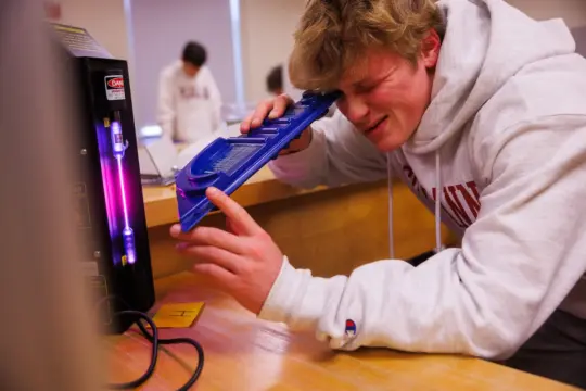 A person wearing a hoodie is squinting while closely examining a mercury spectrum tube on a machine. They're using a blue diffraction grating to view the spectral lines in an educational setting.
