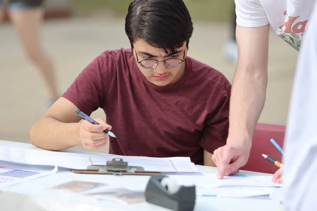 A person wearing glasses and a red shirt is seated at a table outdoors, concentrating on paperwork. Their right hand holds a pen, while another person points at the documents on the table. Office supplies, like a clipboard and tape, are visible.
