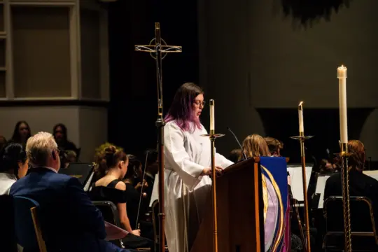 A Pre-Ministry student standing during a church service. Candles are lit beside them, and musicians with instruments are seated nearby. The setting is dimly lit.