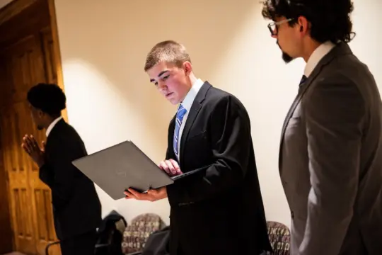 Three people in formal attire are standing indoors. The person in the center is holding and looking at a laptop, while the others are nearby. They are in a hallway with a wooden door in the background.