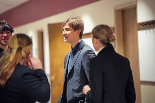A young man in a suit stands in a hallway, smiling while talking to three people. Two women and a man are engaged in conversation with him. The hallway has wooden doors and a brown and white wall.