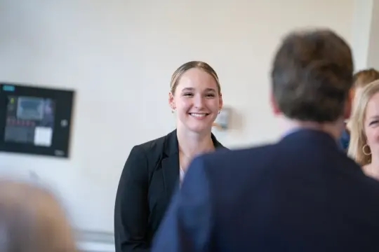 A person in a black blazer is smiling and engaged in conversation with two other people. The setting appears to be indoors, with a blurred background suggesting a professional or social event.