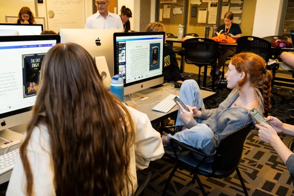 Students in a classroom sit at computers. A red-haired student in a gray sweater casually leans back in their chair, holding a phone. The screens display text and images. Other students are engaged in tasks, while an instructor stands nearby.