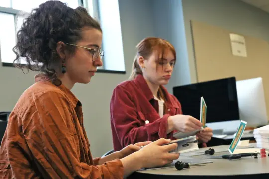 Two people sitting at a table, playing a card game. They appear focused, each holding cards. A computer monitor is visible in the background. Both are wearing casual clothes and glasses. The setting seems to be an indoor room with soft lighting.