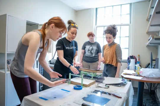 Four people are engaged in a creative project around a table covered with tools and materials. Two are actively working while the others observe. A window is in the background, and shelves are on the right. The atmosphere is collaborative.