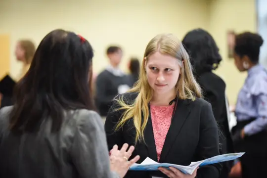 Two women are in conversation at a professional event. The woman on the right is holding a booklet and wearing a black blazer with a pink top. The background is blurred, showing other attendees in discussion.