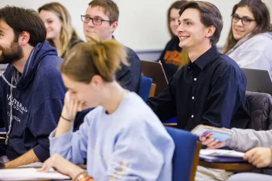 Students sitting in a classroom, listening attentively. A young man in a black shirt smiles while others, wearing casual clothes, focus on their notes and laptops. They are seated at desks, engaging in a lively lecture.