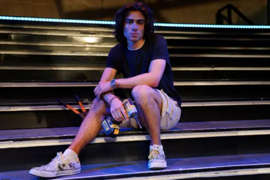 A person with shoulder-length hair sits on steps, holding a cordless drill. They're clad in a black t-shirt, beige shorts, and white sneakers. The outdoor night setting, bathed in blue overhead lighting, resembles an impromptu stage—a scene any Theatre Acting BFA would admire—with tools scattered beside them.