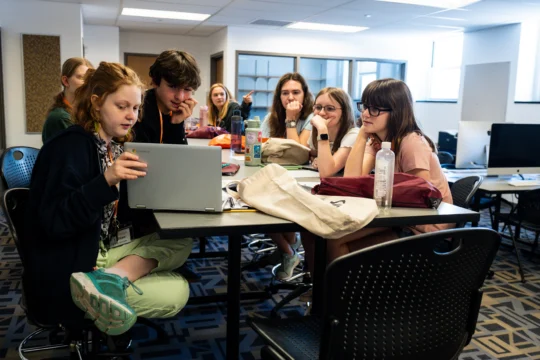 A group of students sit around a table in a classroom, closely looking at a laptop together as they work on publishing & editing. Papers, bags, and water bottles are on the table, with more students and desks in the background.