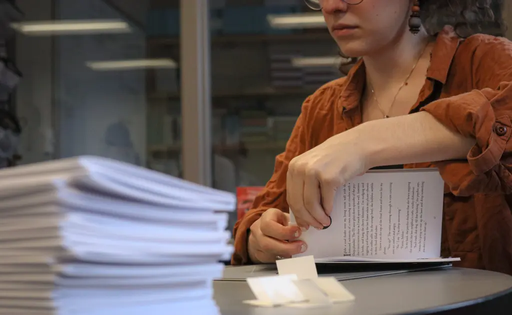A person in a brown shirt is carefully folding a piece of paper at a desk with a stack of papers nearby. With glasses and curly hair, they are fully focused, perhaps pondering English Literature. Shelves brimming with books create an intellectual backdrop.