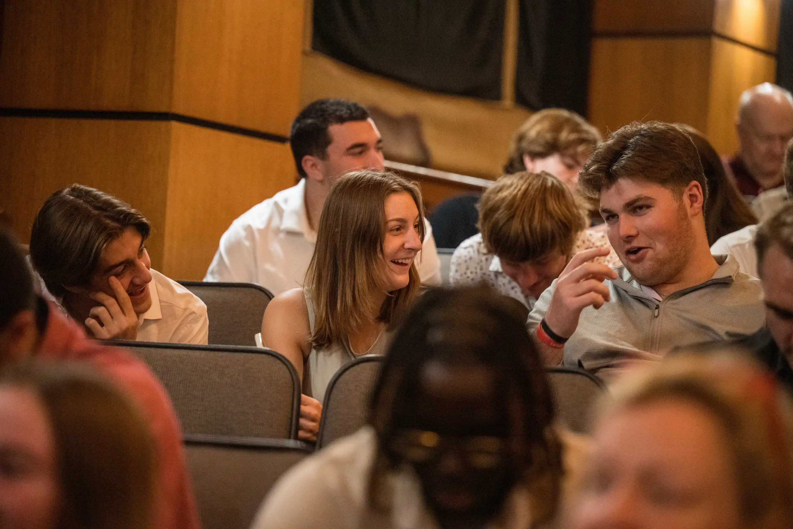 A group of people sit in an auditorium, engaged in conversation and smiling. The atmosphere feels casual and friendly as they discuss philosophy, exchanging ideas and chatting before the event begins.