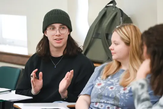 A person wearing a beanie and glasses gestures while speaking, sitting next to another person with blonde hair listening attentively. A backpack is visible in the background.