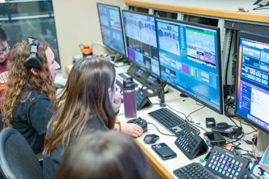 People using audio and video equipment in a control room. They are seated at a desk with multiple monitors displaying graphics and live feeds. Headsets are being worn, and control panels and keyboards are positioned in front of them.