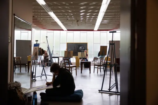 A person sits on the floor sketching in an art studio filled with easels and chairs. The room is well-lit with natural light from the large windows, creating a calm and focused atmosphere. Various artworks are displayed on easels.