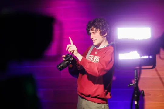 A person with curly hair wearing a red sweater holds a camera and gestures with one hand. They stand in front of a wooden background with purple and white lighting. A bright studio light is visible on the right.