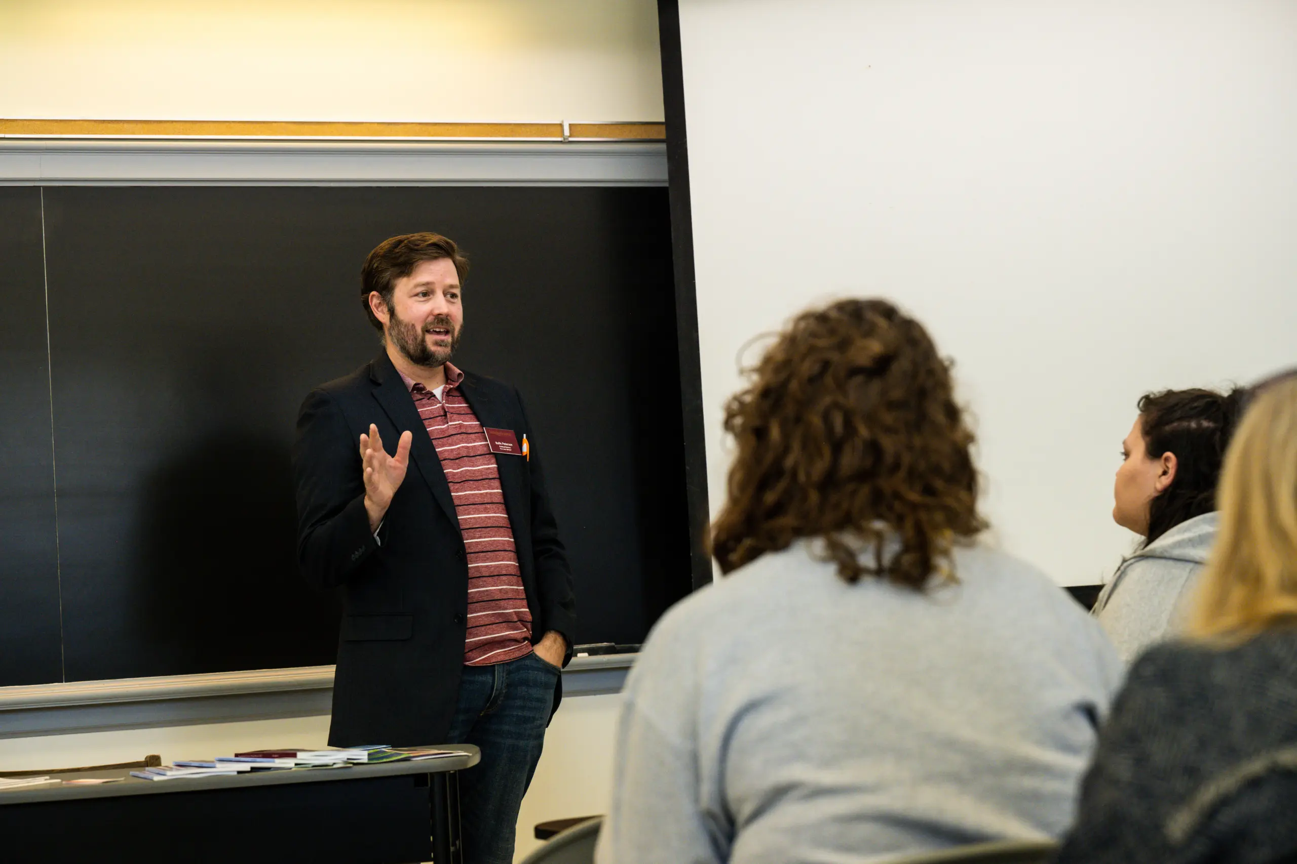 A man with a beard, wearing a blazer and red striped shirt, stands in front of a blackboard and gestures while speaking to a group of people during a Pre-Law Advising session in a classroom.