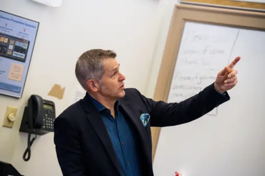 A man in a dark blazer gestures and points while speaking in a classroom, standing near a whiteboard with handwritten history notes and a wall phone in the background.