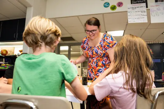 A teacher wearing glasses and a colorful dress smiles and interacts with two young students seated at a table in a classroom. The students, one with blond hair and another with brown hair, focus on their work. Classroom posters are visible in the background.