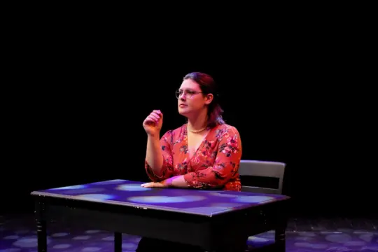 A person with glasses and wearing a red floral blouse sits at a table on a dimly lit stage, gesturing with one hand. The background is dark with spotlight patterns on the table.