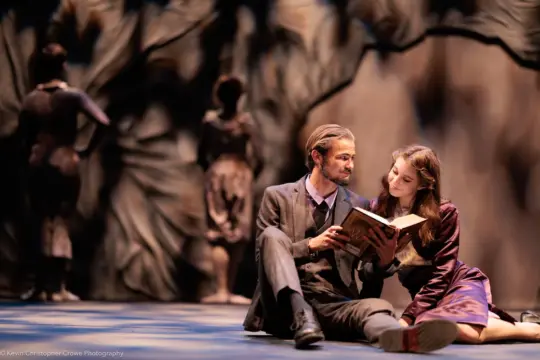 A man and a woman sit on stage, reading a book together. They are dressed in early 20th-century attire. The background features soft, abstract shapes resembling trees and two blurred figures. The lighting is warm and theatrical.
