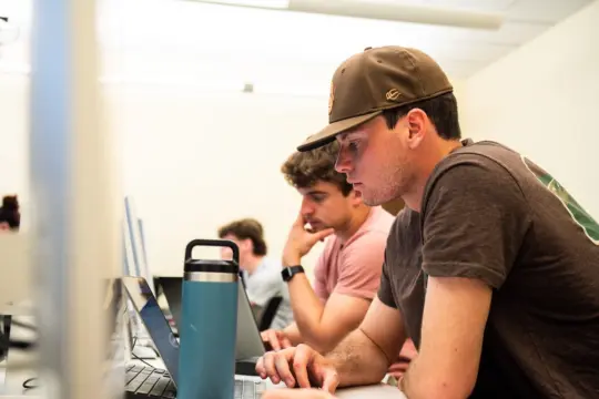 Two young men focus intently on their laptops in a bright room with a white wall. One wears a brown T-shirt and cap, while the other wears a pink T-shirt. A blue water bottle sits on the desk in the foreground.