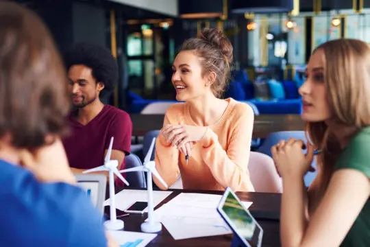 A group of young adults sitting at a table in a modern office, discussing plans. There are small wind turbine models and documents on the table. The atmosphere is collaborative and focused.
