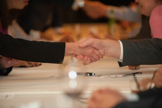 Two people in business attire shake hands across a conference table, with papers, pens, and other blurred individuals in the background, highlighting a Professional Sales meeting or formal business negotiation.
