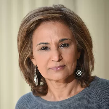 A person with shoulder-length brown hair is wearing a grey sweater and ornate, circular earrings. They are standing against a neutral background.