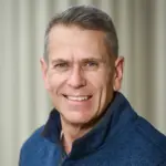 A man with short gray hair smiling warmly, wearing a blue jacket, against a softly blurred background.