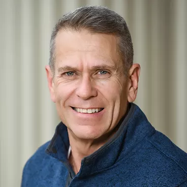A man with short gray hair smiling warmly, wearing a blue jacket, against a softly blurred background.