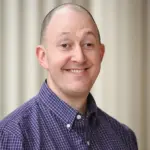 A smiling person wearing a purple checkered shirt stands in front of a light-colored, blurred background.