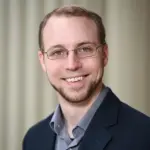 A man with short light hair and glasses, wearing a dark blazer and a gray shirt, smiles at the camera against a neutral background.