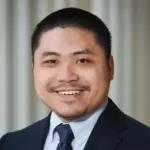 Smiling person in a suit and tie against a neutral background.