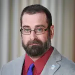 A man with glasses, a beard, and short hair wears a gray suit, red shirt, and blue tie. A pin with a blue and white logo is on his lapel. The background is softly blurred with vertical lines.