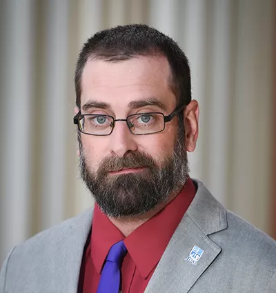 A man with glasses, a beard, and short hair wears a gray suit, red shirt, and blue tie. A pin with a blue and white logo is on his lapel. The background is softly blurred with vertical lines.
