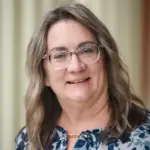 A person with long wavy hair and glasses smiles at the camera, wearing a floral patterned blouse and a gold necklace. The background is softly blurred.