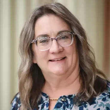 A person with long wavy hair and glasses smiles at the camera, wearing a floral patterned blouse and a gold necklace. The background is softly blurred.