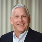 An older man with short gray hair smiles at the camera. He is wearing a dark blazer over a light-colored shirt. The background consists of vertical, light-colored blinds.