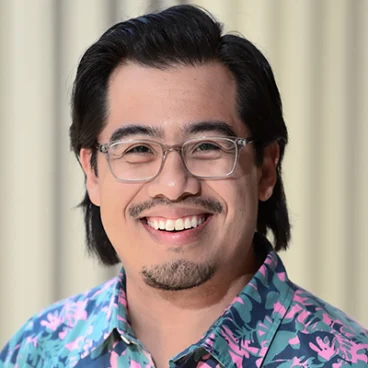 A person with long dark hair, glasses, and facial hair is smiling. They are wearing a blue shirt with a colorful pattern. The background shows vertical blinds in soft focus.